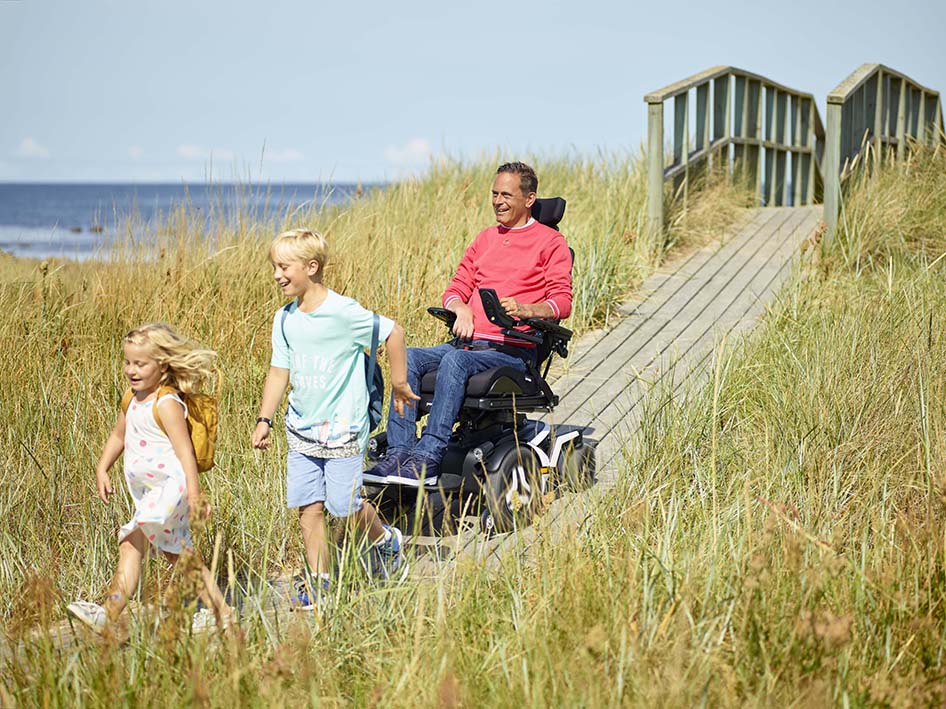 Man driving Permobil F5 Corpus towards beach area with children 